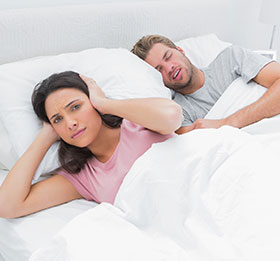 young woman in bed holding her hands over her ears while her husband snores